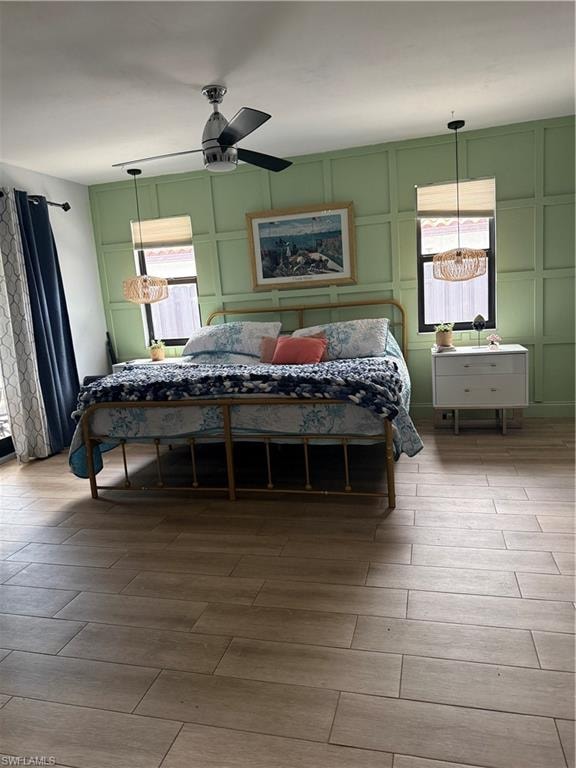 Room featuring wood-finish flooring, a decorative accent wall, a ceiling fan, and two windows with pendant light fixtures
