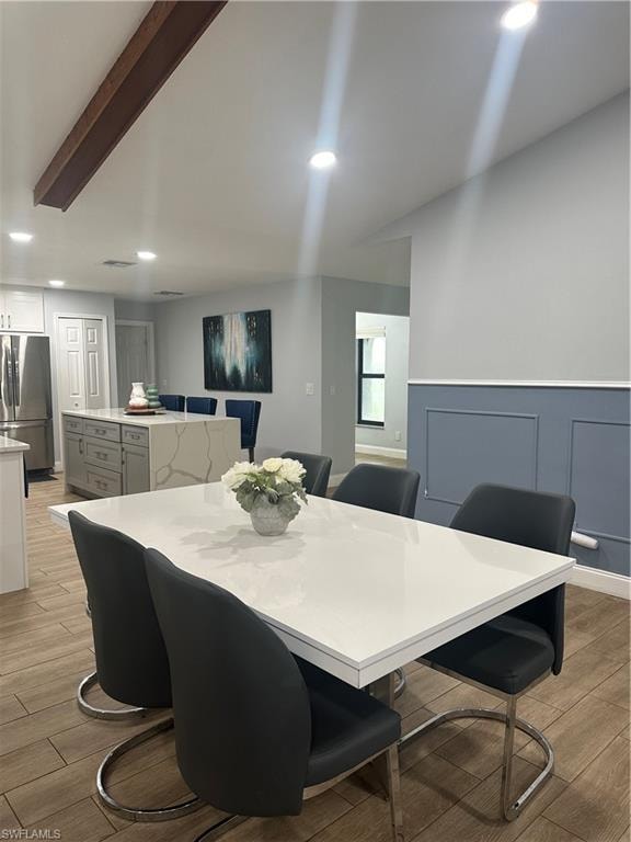 Open-concept dining area featuring wood-finish flooring, recessed lighting, a decorative ceiling beam, a white countertop dining table, and wainscoting
