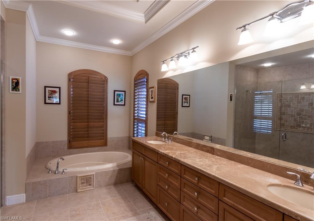 Dual vanity with stone countertop and integrated sinks, a built-in tub with tile surround, a large frameless glass shower with mosaic tile detailing, architectural arched window shutters, and recessed lighting