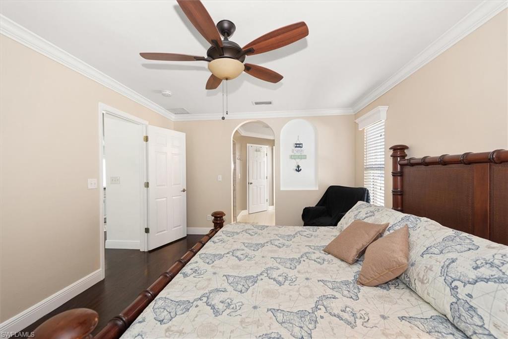 Spacious bedroom featuring wood-finish flooring, crown molding, a ceiling fan with light fixture, arched doorways, and a window with horizontal blinds