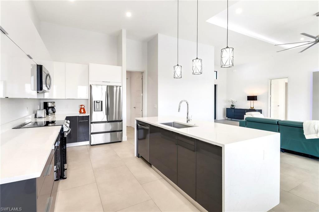 Open-concept kitchen featuring an oversized island with an undermount sink and three pendant lights, stainless steel appliances, white cabinetry, and light-toned tile flooring