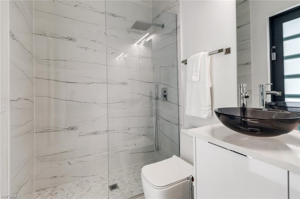 Contemporary bathroom featuring a clear glass shower enclosure with a rainfall shower head, white marble-style wall tiling, pebble-finish shower pan, black vessel sink, and a white vanity with a light-toned countertop