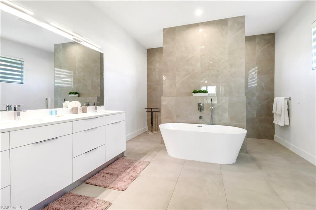 Spacious bathroom featuring a freestanding soaking tub with a wall-mounted faucet, a double vanity with ample storage, large format floor tiling, a full-width mirror, and an accent wall with large format stone-finish tiles