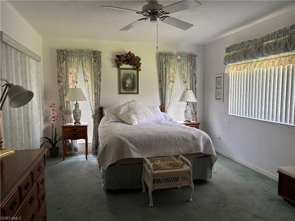 White walls with a ceiling fan, light blue carpeting, and windows featuring vertical blinds and floral valances