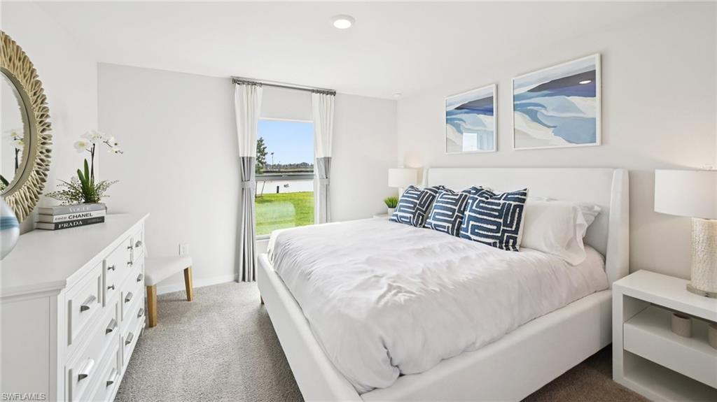 Model Home. This bedroom features light-colored walls and carpeting, a window with a view of a body of water and green space, and a white dresser with a round mirror