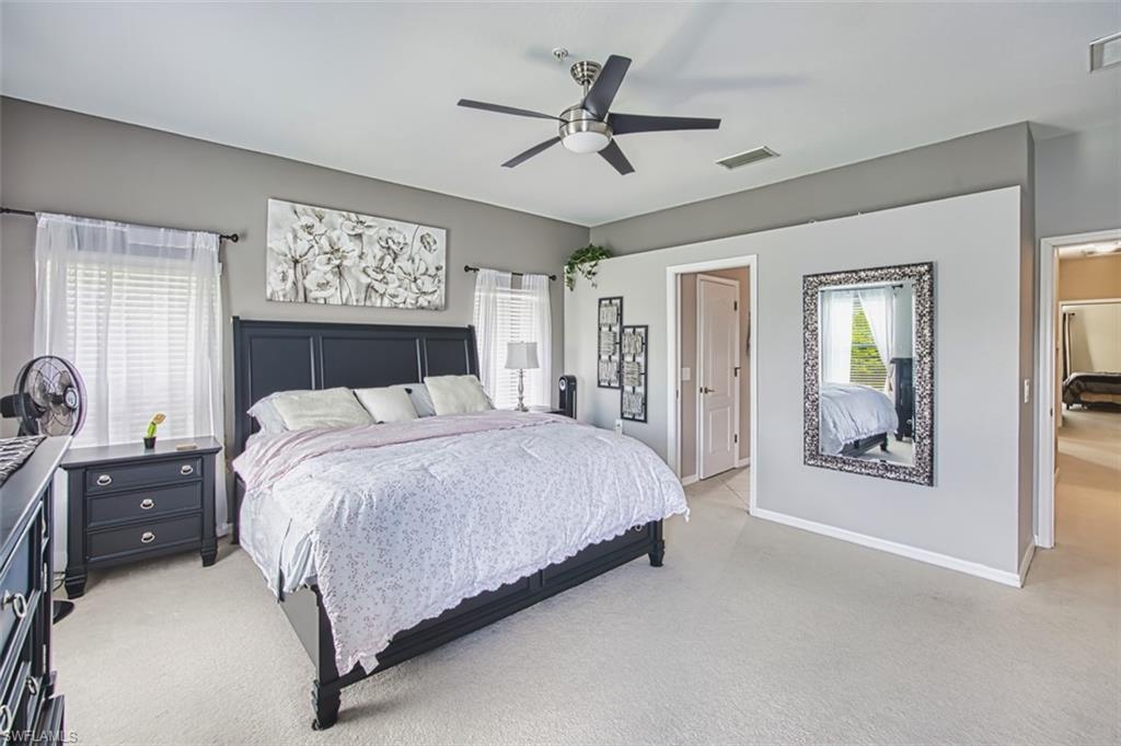 Spacious bedroom featuring light-toned carpeting, neutral wall colors, dual windows with blinds and sheer curtains, a ceiling fan with integrated lighting, and multiple entryways