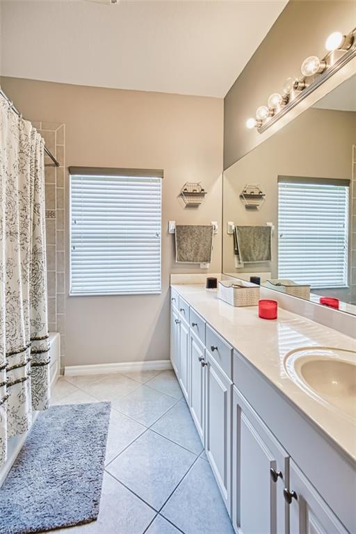 Bathroom featuring a dual-sink vanity with white cabinetry and a light-toned countertop, a large mirror with overhead vanity lighting, a window with horizontal blinds, and tile flooring