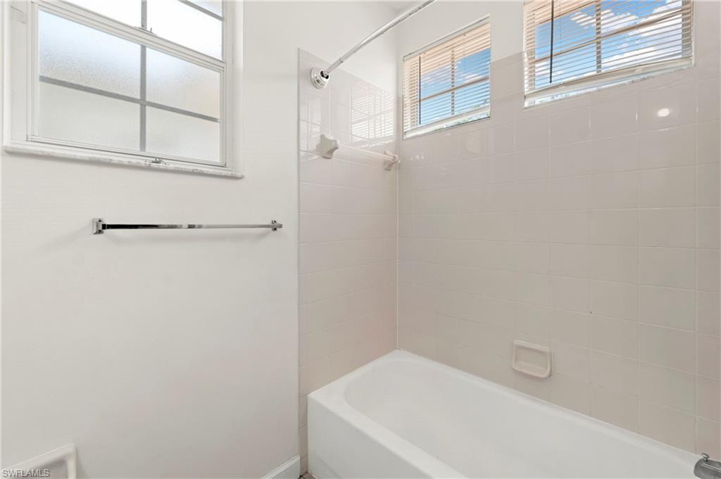 Bathroom with a white tub and shower combination, featuring white tile surround, two windows with horizontal blinds, and a frosted glass window