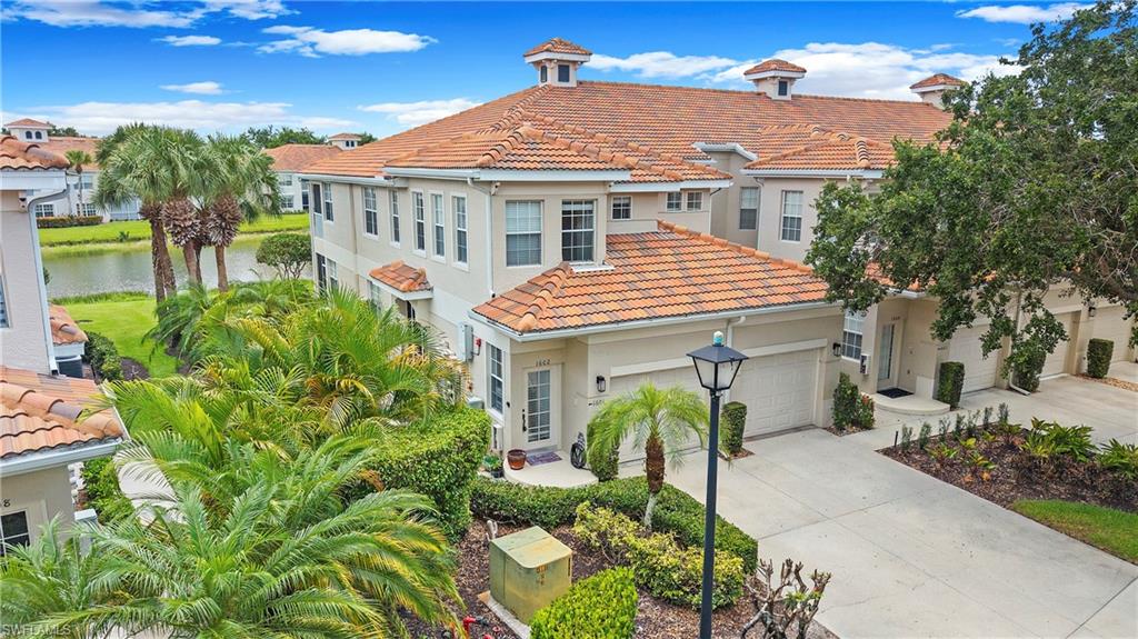 Stucco exterior featuring a terracotta tile roof, attached garage, paved driveway, and established tropical landscaping