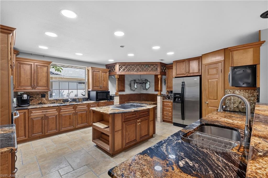 Expansive kitchen featuring wood cabinetry, varied granite countertops, a stainless steel refrigerator, a center island with cooktop, and recessed lighting