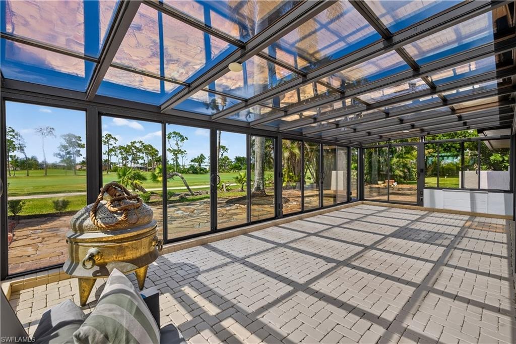 Spacious sunroom featuring a clear glass and metal frame ceiling, floor-to-ceiling glass panel walls, light-toned paver flooring, and white built-in cabinetry