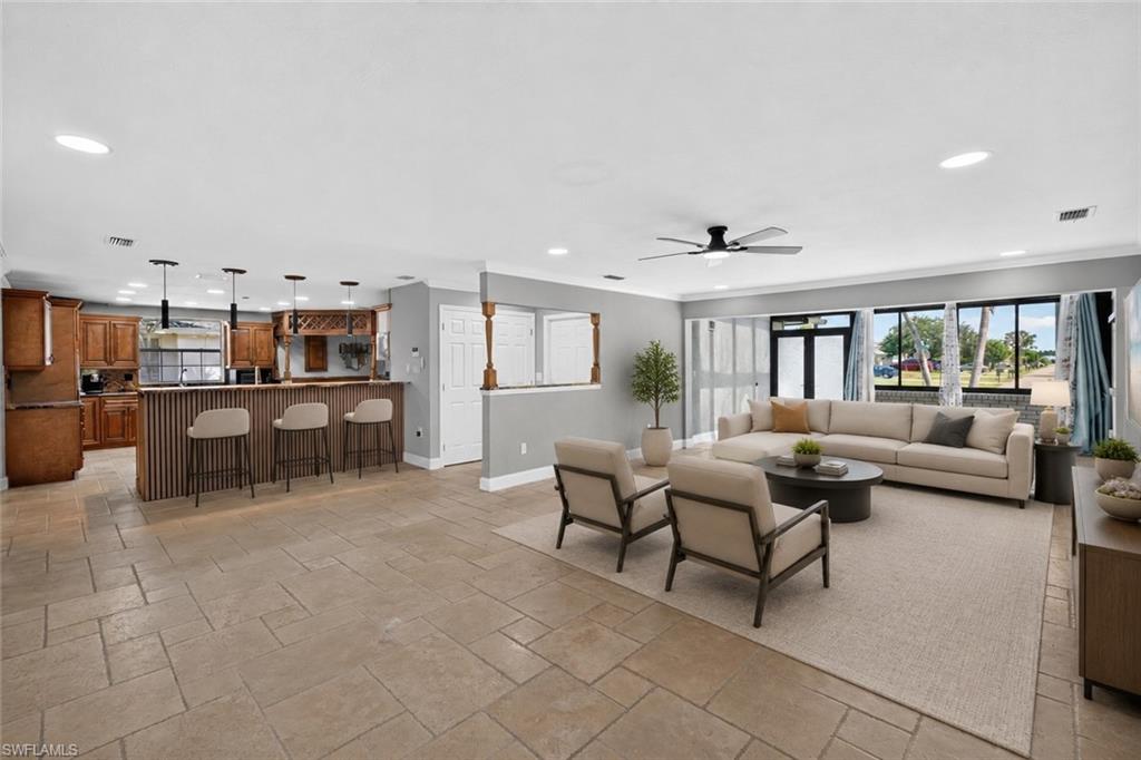Open concept living area featuring light-toned tile flooring throughout, a kitchen with wood cabinetry, and a living room with multiple large windows, Virtually Staged