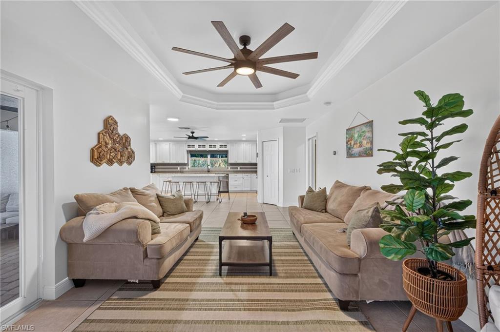 Spacious open-concept living area featuring a tray ceiling with a multi-blade ceiling fan, crown molding, and light-toned tile flooring