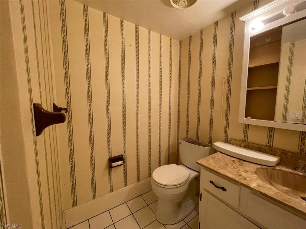 Bathroom featuring a white toilet, white vanity with integrated sink, patterned wallpaper, white tile flooring, and wall-mounted lighting fixture