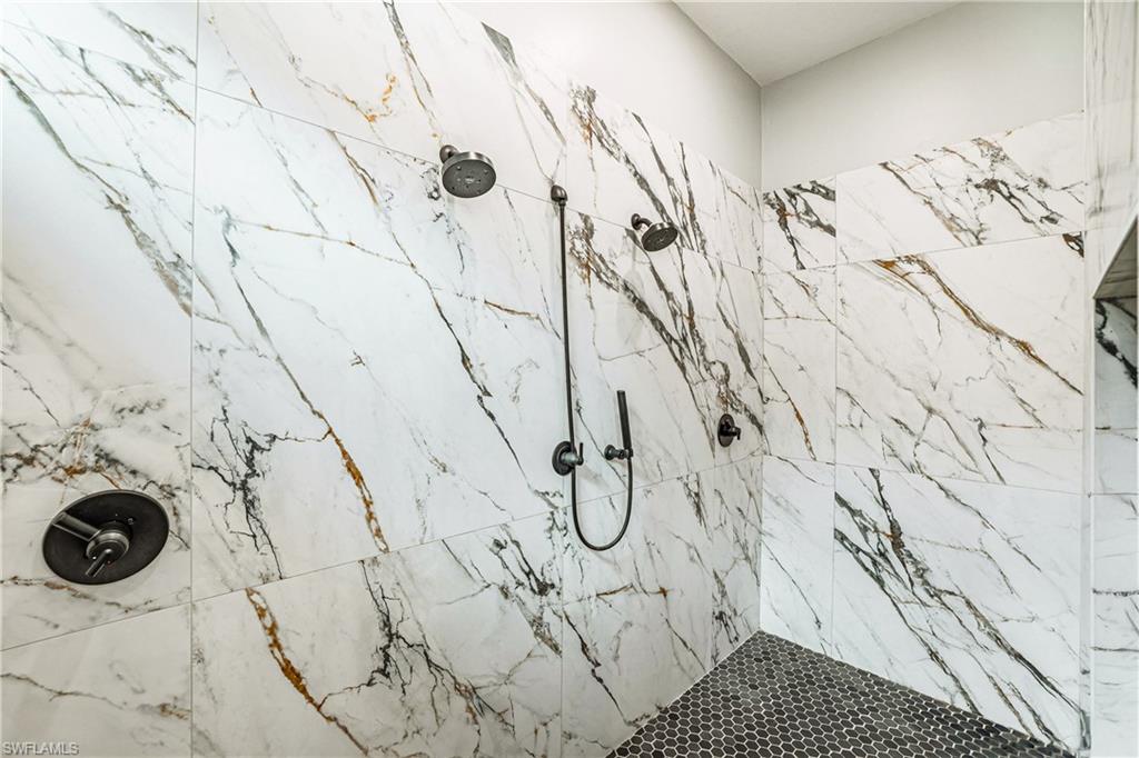 Spacious shower with large-format marble-look wall tiling, dual shower heads, a handheld shower fixture, and dark hexagonal floor tiling