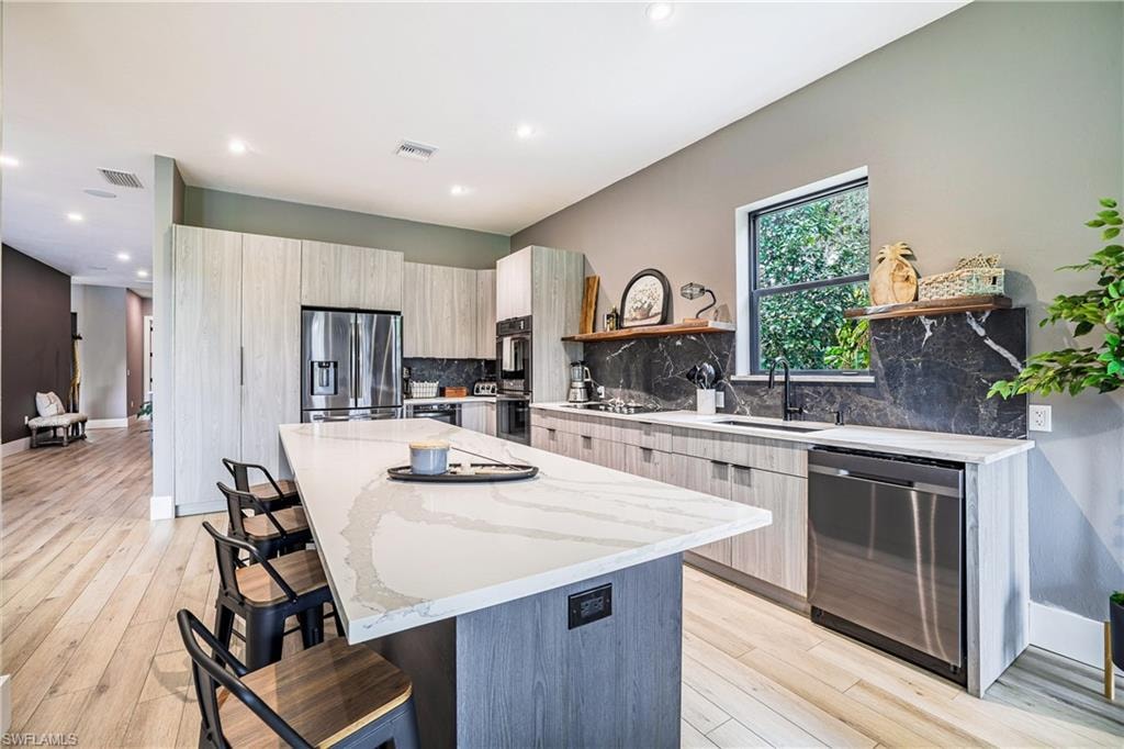 Contemporary kitchen featuring a large central island with seating, light wood-finish flooring, full-height cabinetry, stainless steel appliances, and a dark stone backsplash