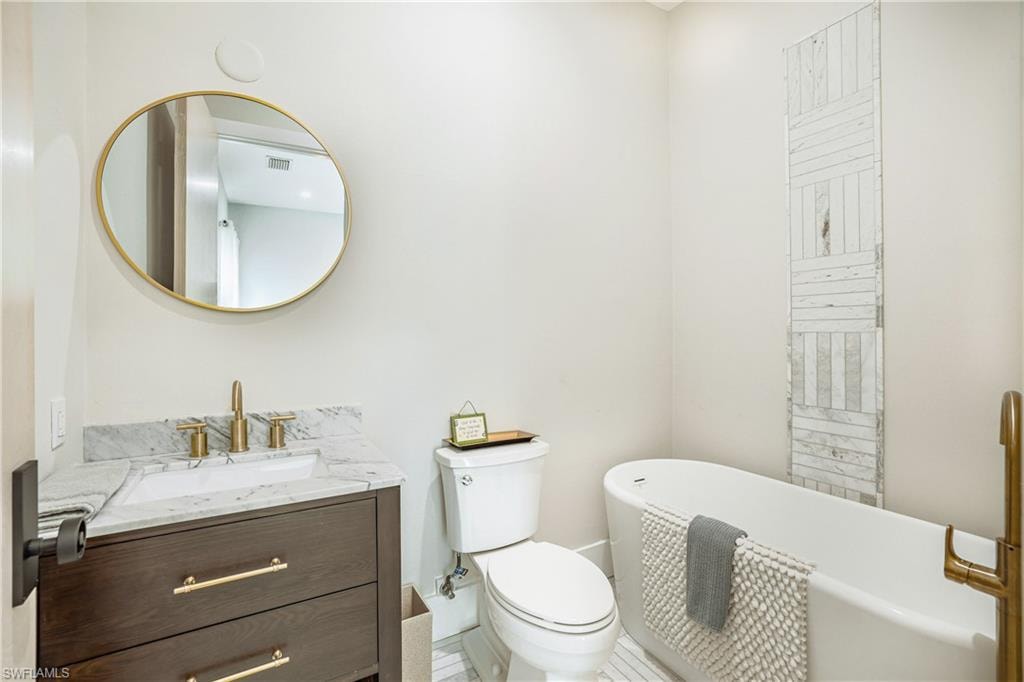 Freestanding soaking tub with a floor-mounted brass faucet, marble-patterned tile accent wall, dark wood-finish vanity with brass hardware, undermount sink, and a circular brass-framed mirror