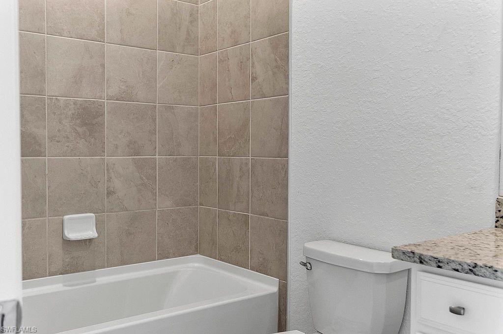 Bathroom featuring a white bathtub, neutral-toned tiled shower surround, textured white walls, a white toilet, and a vanity with a light-colored countertop