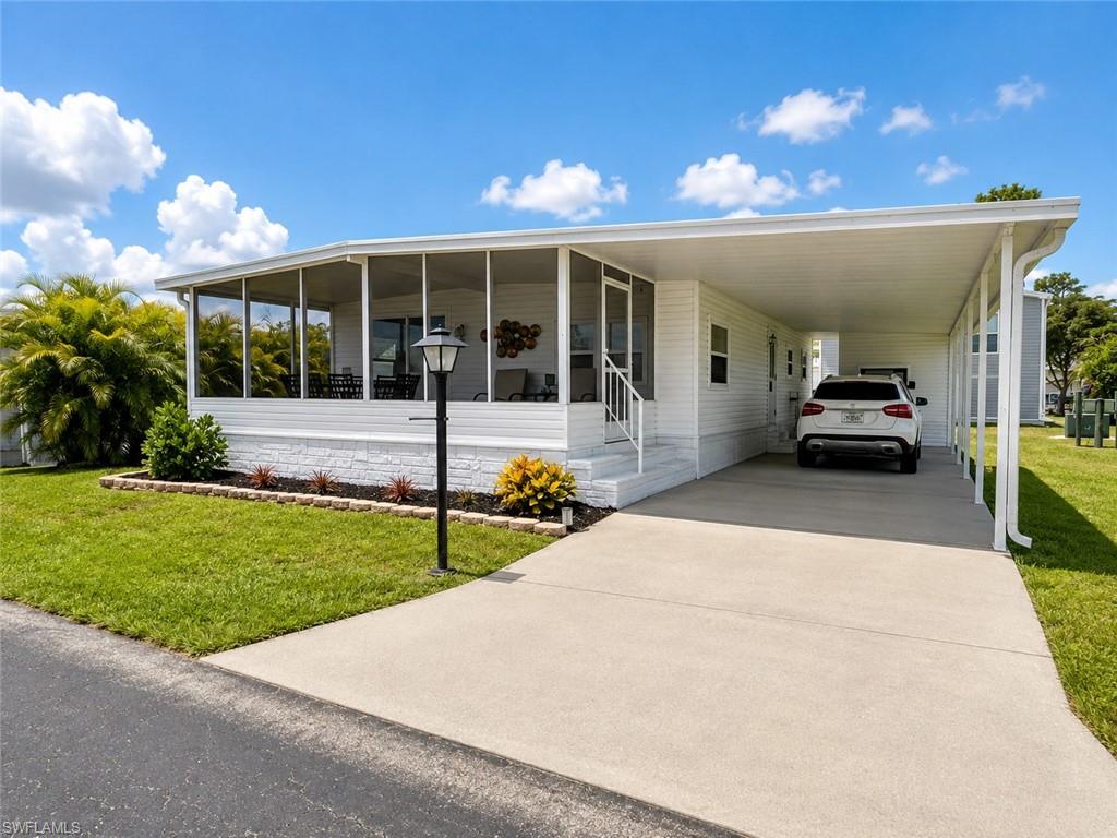 Modern mobile home featuring a covered front porch with large windows, attached carport with concrete driveway, white exterior siding, manicured lawn with mature landscaping, and a street-level approach with minimal grade transition
