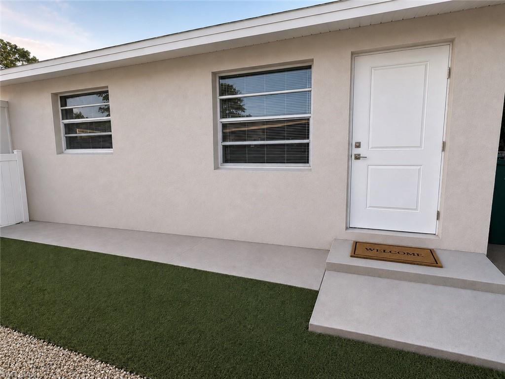 Light-toned stucco exterior featuring a white paneled door with brushed nickel hardware, two windows with horizontal blinds, and a concrete patio slab