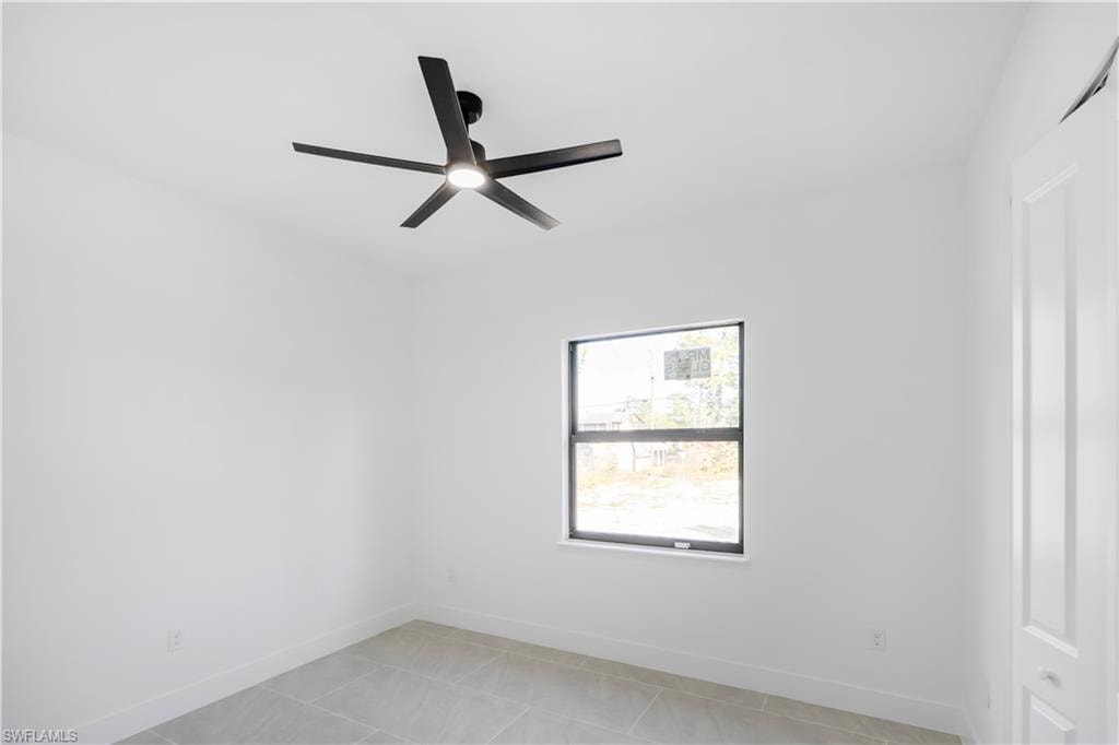 Bright interior room featuring a modern black ceiling fan with integrated lighting, a single pane window with dark trim, light-toned tile flooring, white walls, and white baseboards