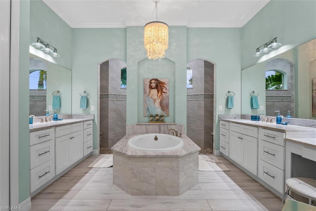 Hexagonal soaking tub with stone surround, dual vanities featuring white cabinetry and light countertops, two arched walk-in showers with tiled walls, wood-finish flooring, and a decorative chandelier