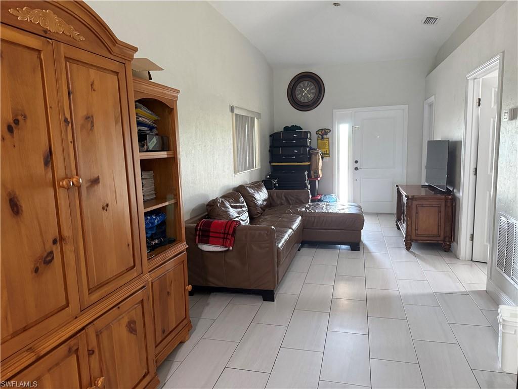 Bright living area featuring vaulted ceilings, light-toned rectangular floor tiles, and a white entry door with a frosted glass insert