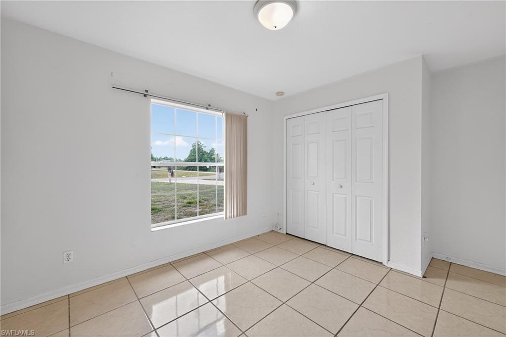 Spacious room featuring neutral tile flooring, a large window with vertical blinds, a ceiling-mounted light fixture, and white bi-fold closet doors