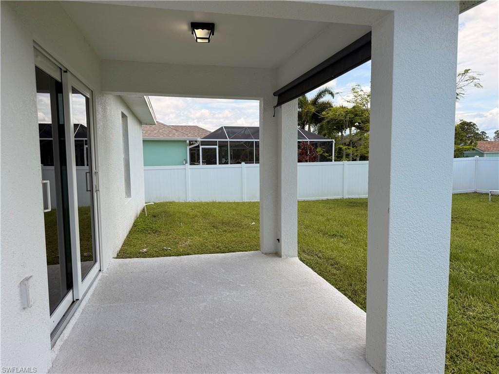 Covered concrete patio with overhead lighting, leading to a fenced backyard featuring a grassy lawn and mature foliage