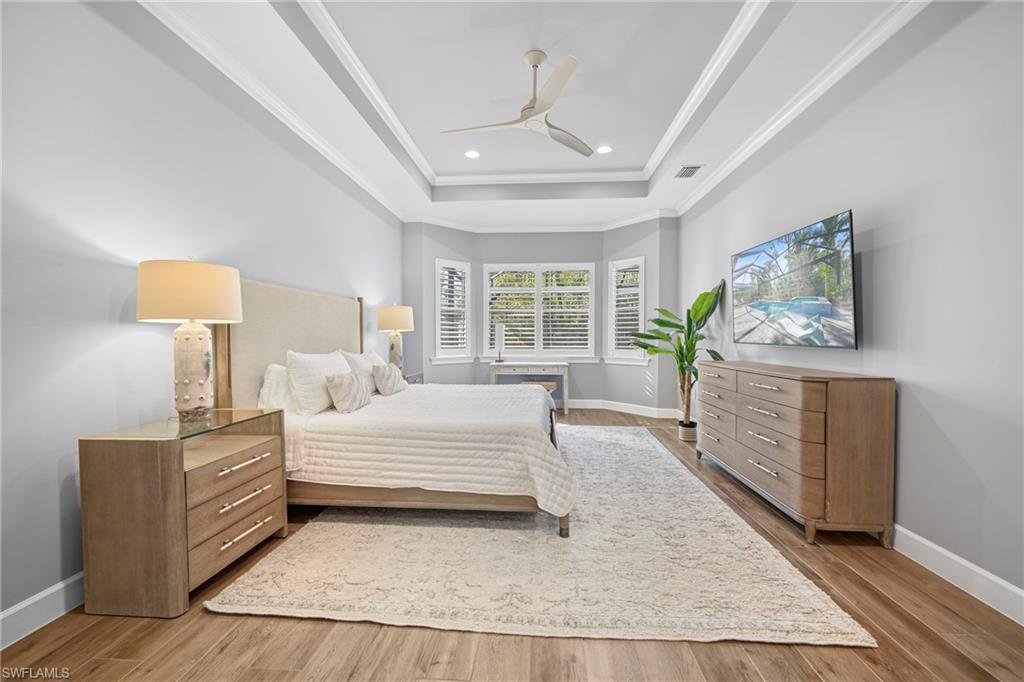 Recessed tray ceiling with crown molding, wood-finish flooring, and a bay window featuring louvered shutters