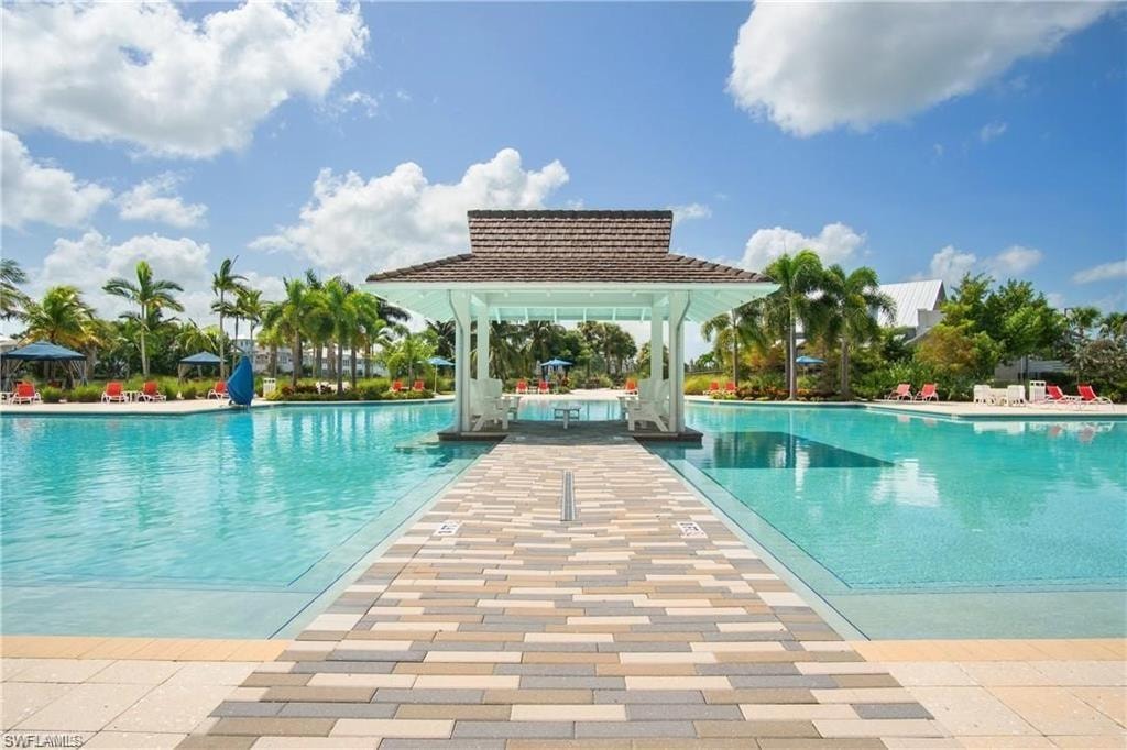 Expansive swimming pool area featuring a central gazebo with a shingled roof, surrounding palm trees, and an extensive paver deck