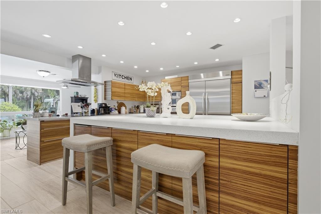 Spacious kitchen featuring a large central island with a light-toned countertop, wood-finish cabinetry, stainless steel appliances, recessed lighting, and a large window providing ample natural light