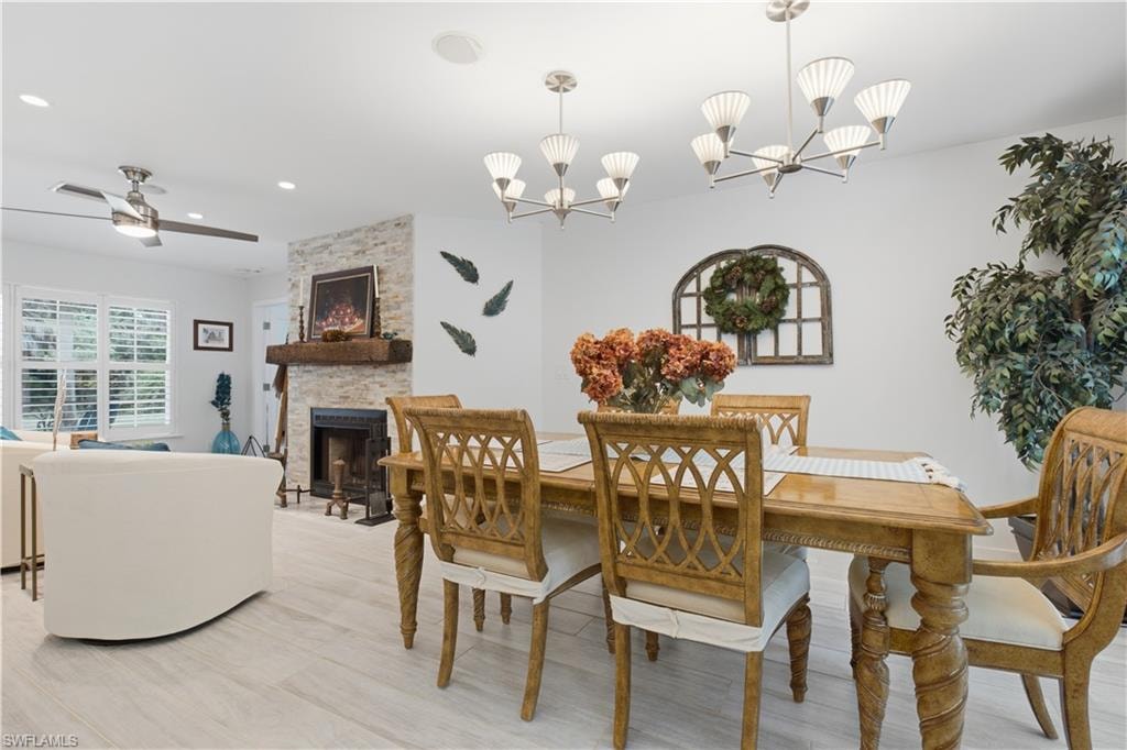 Bright interior space featuring light wood-finish flooring, a stacked stone fireplace with a substantial wood mantel, a ceiling fan with integrated lighting, recessed lighting, and multiple modern chandeliers