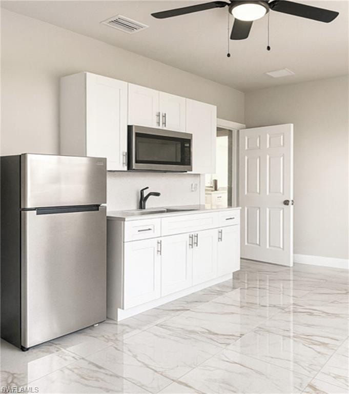 Compact kitchen area featuring white shaker-style cabinetry, stainless steel appliances, a black ceiling fan with integrated lighting, and large format marble-look tile flooring
