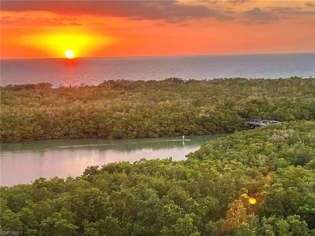 Expansive coastal scenery featuring a vibrant sunset over the ocean, a winding waterway surrounded by lush green foliage, and a distant bridge structure
