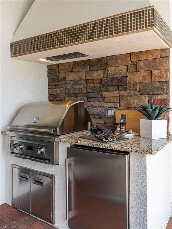Outdoor kitchen featuring stacked stone backsplash, stainless steel grill, under-counter refrigerator, built-in storage, and granite countertop