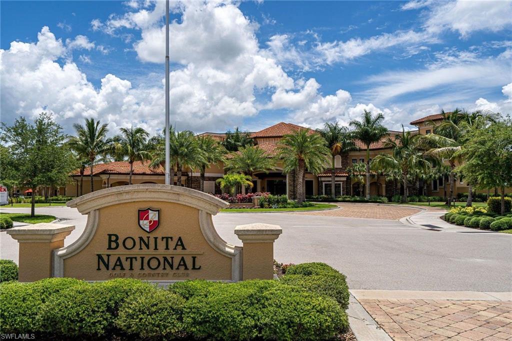 Gated community entrance with an architectural sign, lush landscaping, and paver driveways leading to a large building with a terracotta tile roof and extensive palm trees