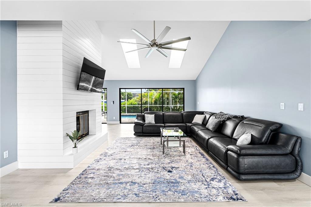 Living room with high vaulted ceiling, a large fireplace, light wood finished floors, a skylight, and a ceiling fan