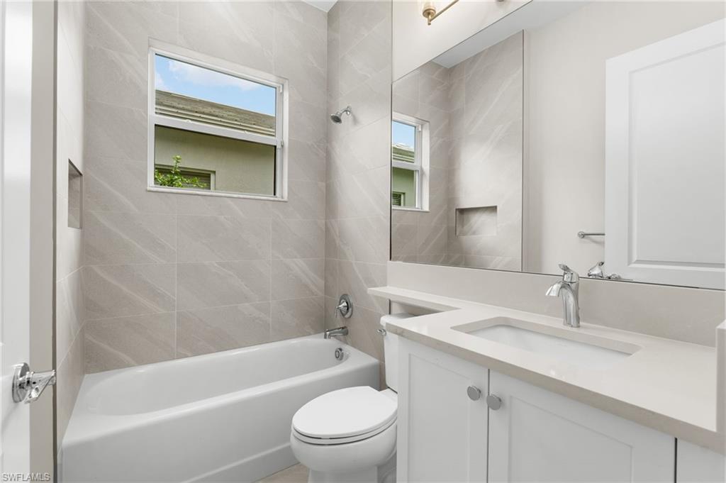 Full bathroom featuring a single vanity with an integrated sink, quartz banjo style countertop a large mirror, a built-in bathtub with shower, full wall tiling