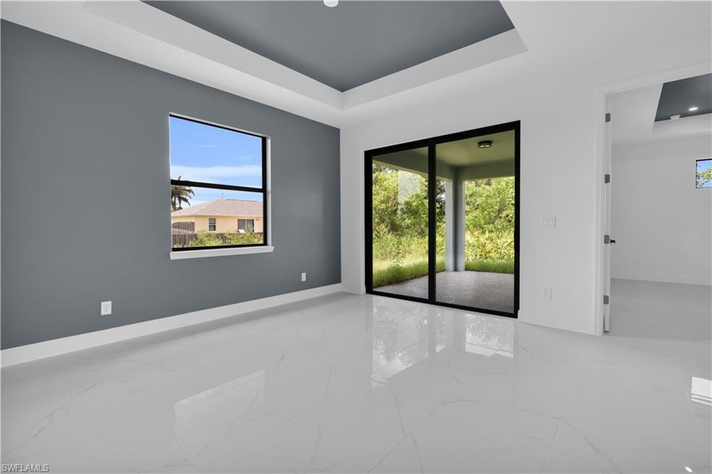 Spacious interior featuring a recessed tray ceiling, large format tile flooring, a window with outdoor scenery, and sliding glass doors opening to a covered patio