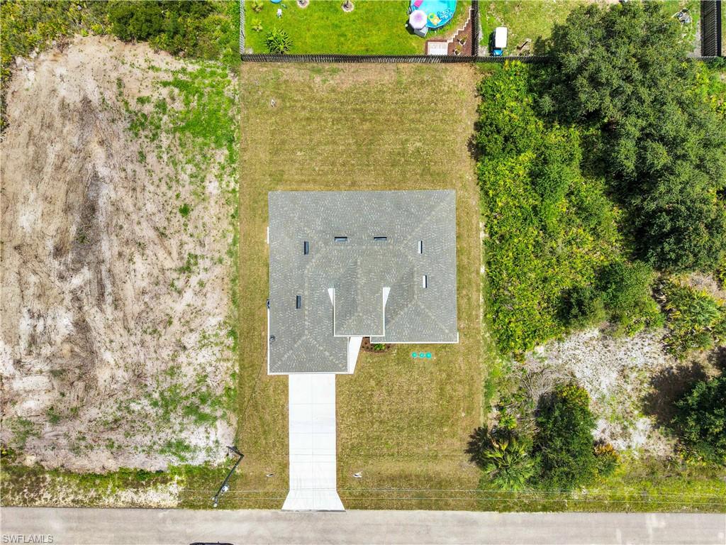 Aerial view of a property featuring a single-story residence with a gray shingle roof, a concrete driveway, and surrounding green lawn