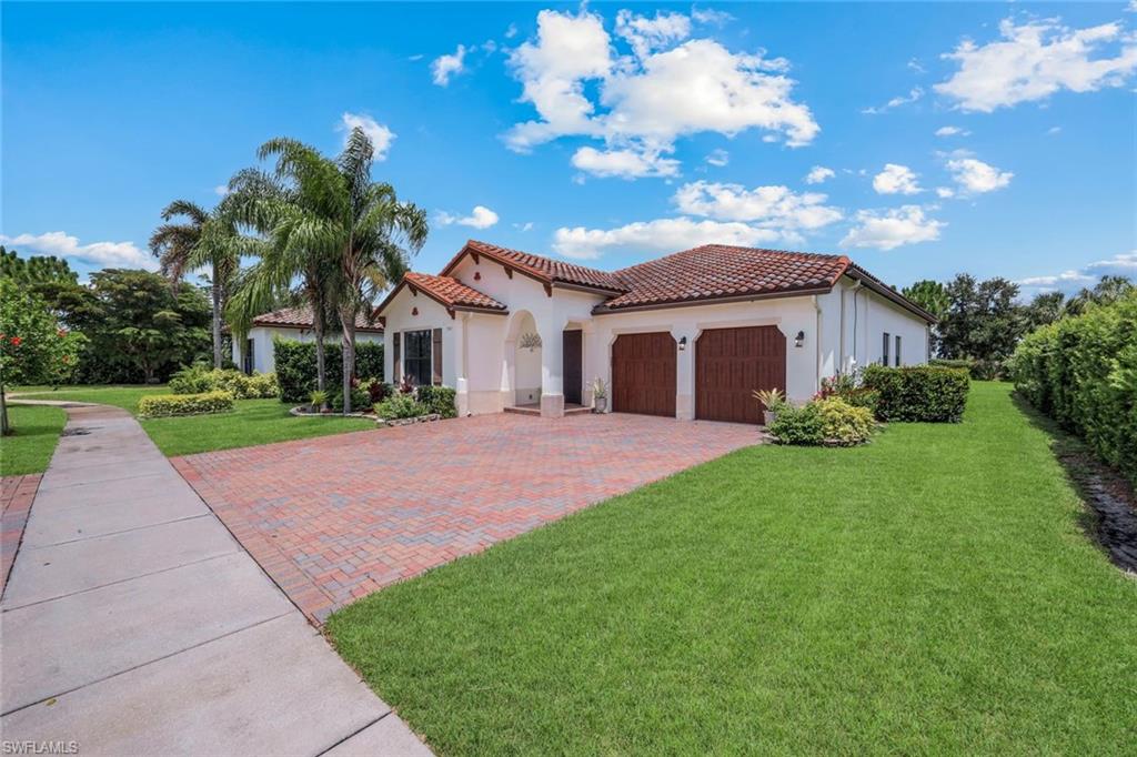 Mediterranean-style exterior featuring a red tile roof, stucco finish, wood-look garage doors, a brick paver driveway, and mature landscaping
