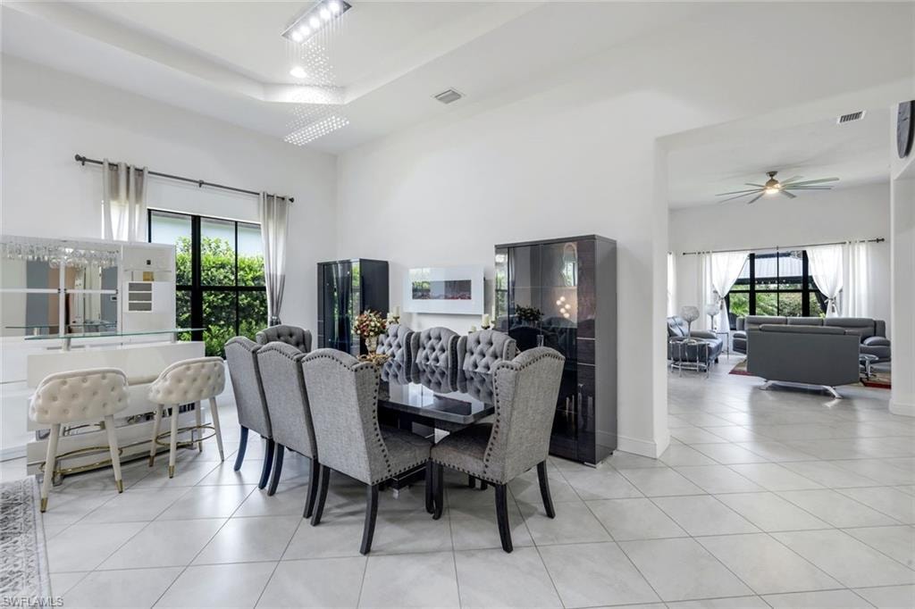 Spacious dining area featuring neutral-toned tile flooring, tray ceiling with recessed lighting, and a built-in bar with overhead glass rack