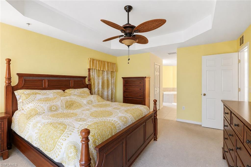 Virtually Staged Carpeted bedroom featuring a tray ceiling with a ceiling fan, light yellow painted walls, and a doorway opening to a tiled area with a built-in tub