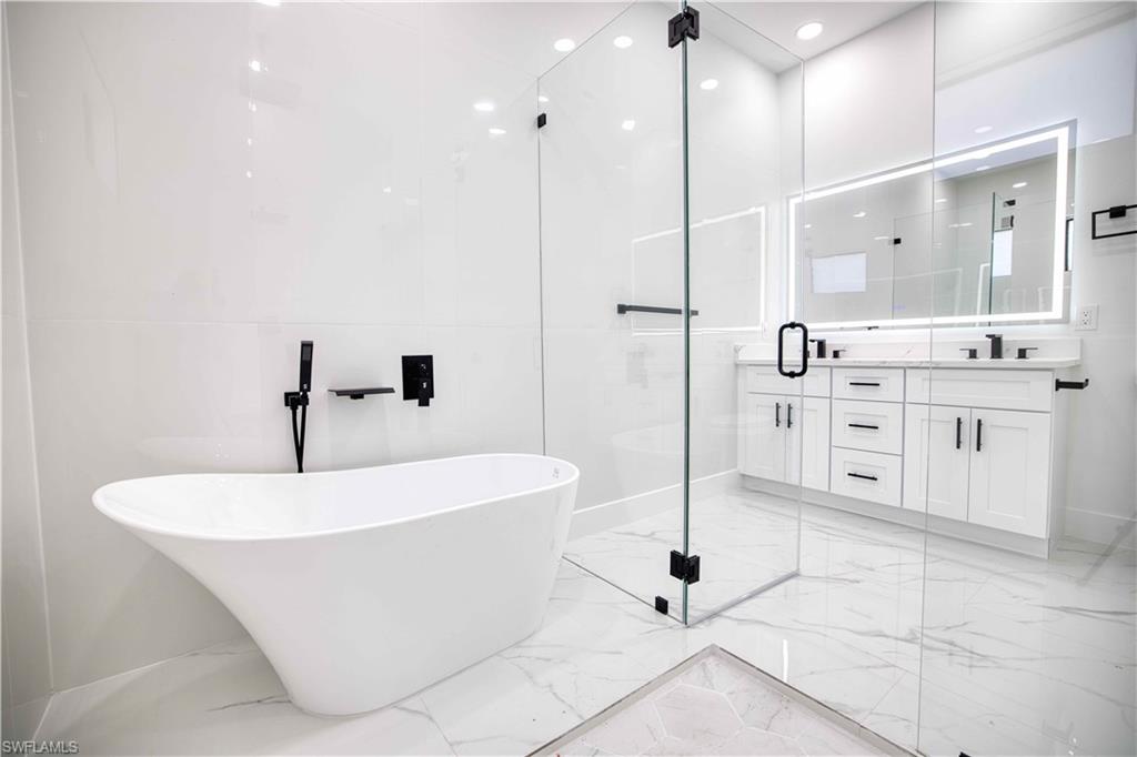 Freestanding soaking tub with wall-mounted matte black faucet, glass-enclosed shower, dual vanity with shaker-style cabinetry, quartz countertop, and illuminated mirror
