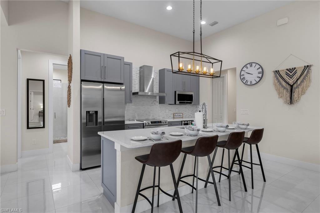 Kitchen with wall chimney range hood, gray cabinetry, a kitchen island with sink, and appliances with stainless steel finishes