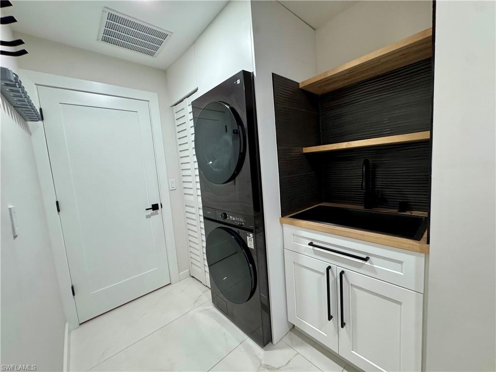 Washroom featuring stacked washer / drying machine, light marble finish floors, and cabinet space