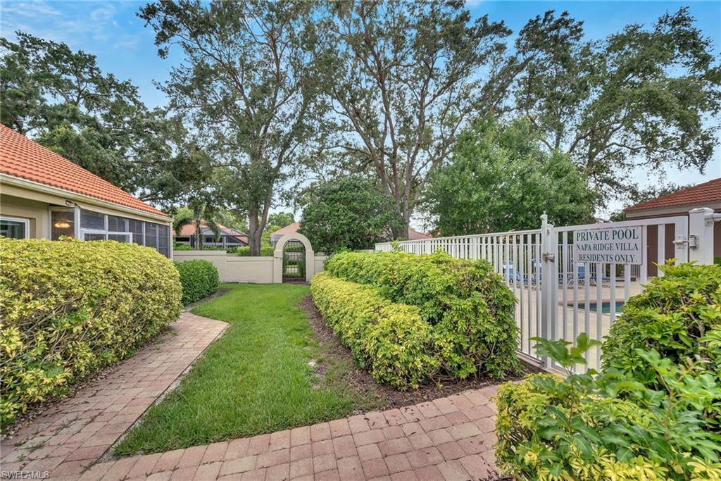 Brick paver pathway with manicured hedges leading to a private pool area, featuring a white fence and lush landscaping