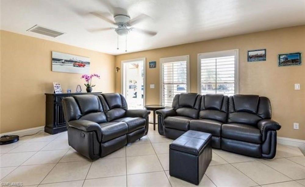 Spacious living area featuring light-toned tiled flooring, neutral wall tones, multiple windows with white plantation shutters, and a ceiling fan with integrated lighting