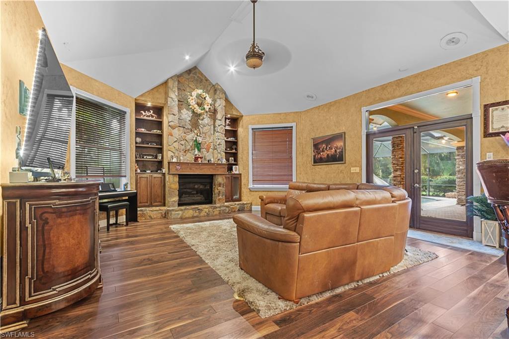 Spacious room featuring vaulted ceilings, wood-finish flooring, a stone fireplace with built-in shelving, and double glass doors